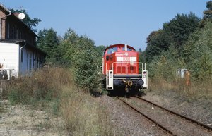 Frontaufnahme der Museumsbahn. Am linken Bildrand sieht man ein Teilstück eines Bahnhofgebäudes, im Hintergrund sind Bäume, Büsche zu sehen.