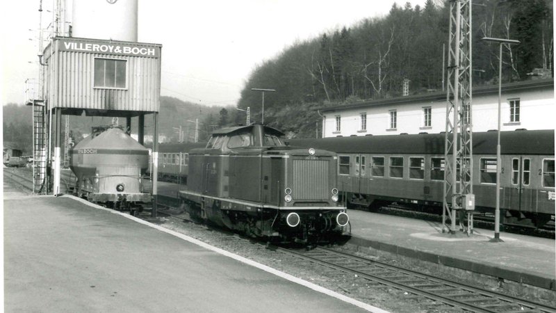 © Bernd Melcher, Sammlung Georg Dollwet schwarz-weiß Fotografie von 1971. Es ist eine Lok im Bahnhofsbereich zu sehen. Auf den Schienen dahinter ist ebenfalls ein Zug mit mehreren Waggons zu sehen.