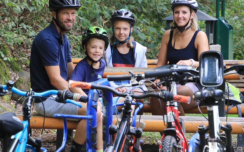 Familie an einer Sitzgruppe. Auf der hinteren Sitzbank sitzen links die Tochter, rechts die Mutter, auf der vorderen Band sitzen links der Vater, rechts der Sohn. Alle 4 Personen tragen Fahrradhelme, die Räder sind im vorderen Bildbereich zu sehen. 