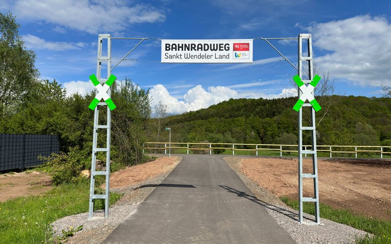 © Nils Veit Das Eingangstor am Bahnradweg besteht aus 2 hohen Eisengestängepfosten, die mit einem Drahtseil verbunden sind. An diesem Drahtseil ist ein Banner befestigt mit der Aufschrift Bahnradweg Sankt Wendeler Land und dem Logo des Weges. An den beiden Eisenpfosten ist jeweils ein grün-weißes Andreaskreuz aufgehängt..