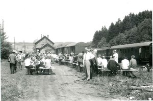 schwarz-weiß Foto einer Feierlichkeit am Bahnhof Nonnweiler 1971. im Vordergrund sind viele Menschen zu sehen, die an Tischen sitzen und sich unterhalten. rechts davon ist ein Zug auf den Gleisen zu sehen. im Hintergrund erkennt man die beiden Giebel der Bahnhofgebäuden. 