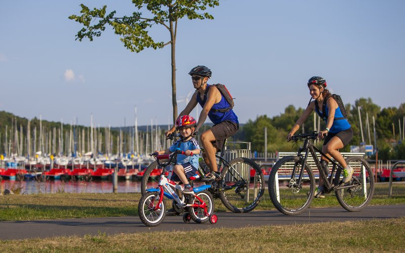 Eine Familie fährt Fahrrad. Ein kleiner Junge fährt vorne, direkt dahinter die Eltern. Im Hintergrund sind der Bostalsee zu sehen mit seinem Hafen, die Mäste der Segelboote sowie der Tretbootsteg mit den roten Tretbooten.