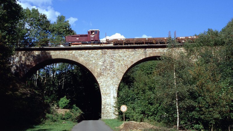 © Marcus Ruch über das Nonnweiler Viadukt fährt die Museumsbahn mit ihrem roten Zug und mehreren Waggons. An den Bildrändern sind Bäume und Büsche zu sehen. Der Himmel ist kaum bewölkt und klar.