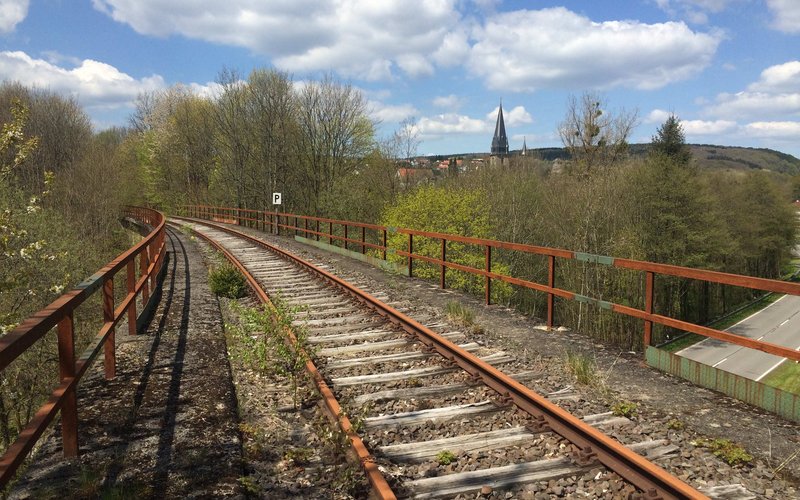 Das Bild zeigt Schienen, die über eine Brücke führen. Im Hintergrund sind Bäume, Sträucher zu sehen. Am oberen Bildrand ist die Turmspitze einer Kirche zu sehen.