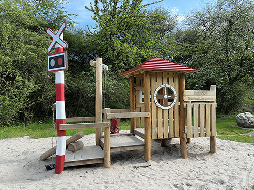 Kinderspielgerät im Bahnhofsdesign. Es gibt unter anderem ein Häuschen, links davon ein Pfosten mit einem Sandeimer, der an einer Eisenkette befestigt ist, eine Fallrinne zum Befördern des Sands